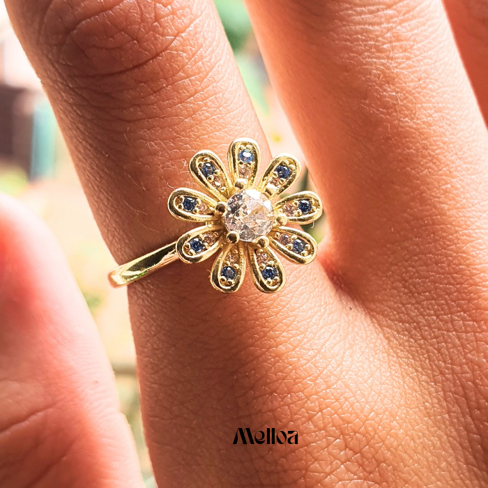 Gold daisy fidget ring with blue stones on the petals and a clear central gem, worn on a hand in natural light, lifestyle photo by Melloa